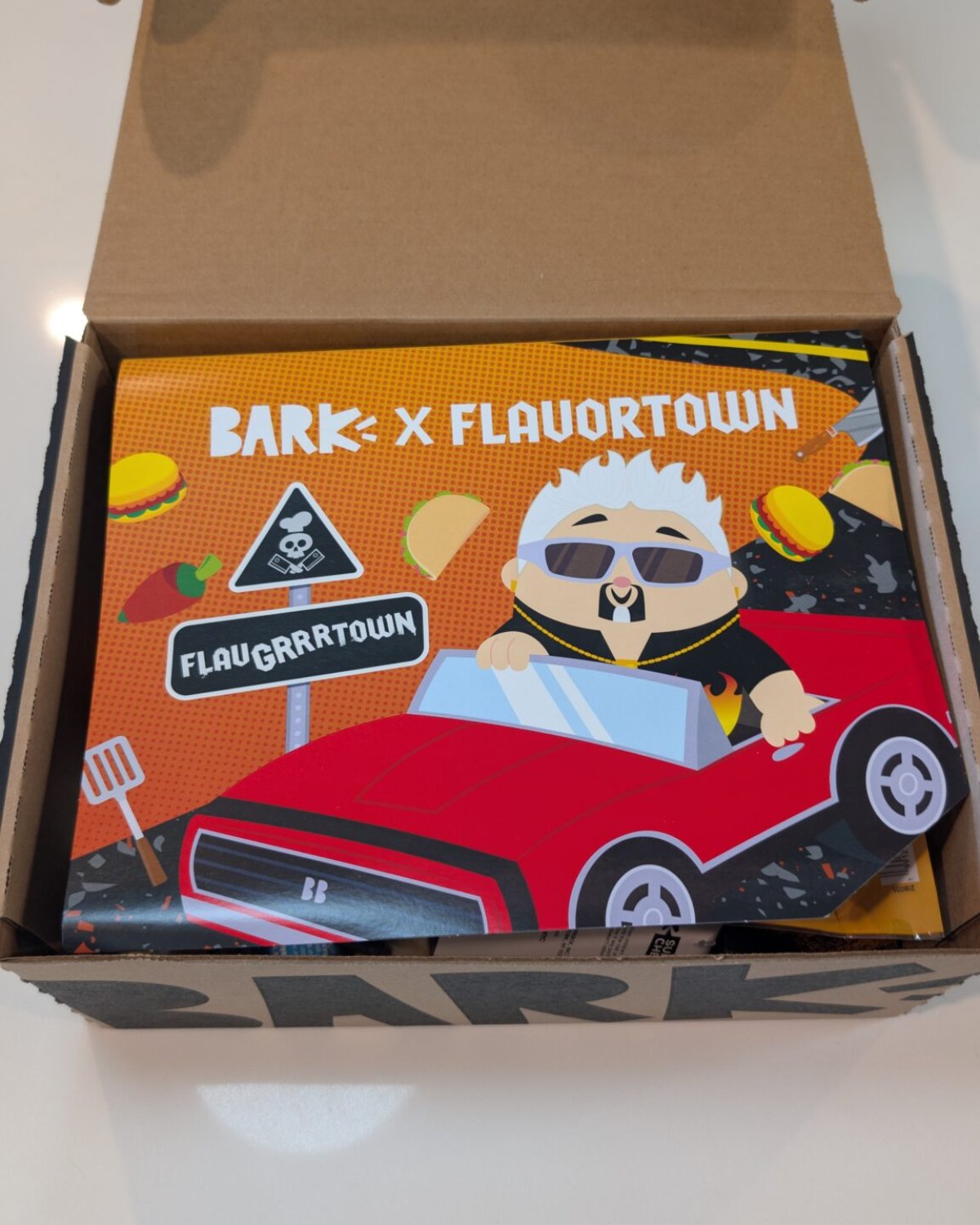 BARK x Flavortown theme card with cartoon Guy Fieri in a red car sitting on top of the opened box