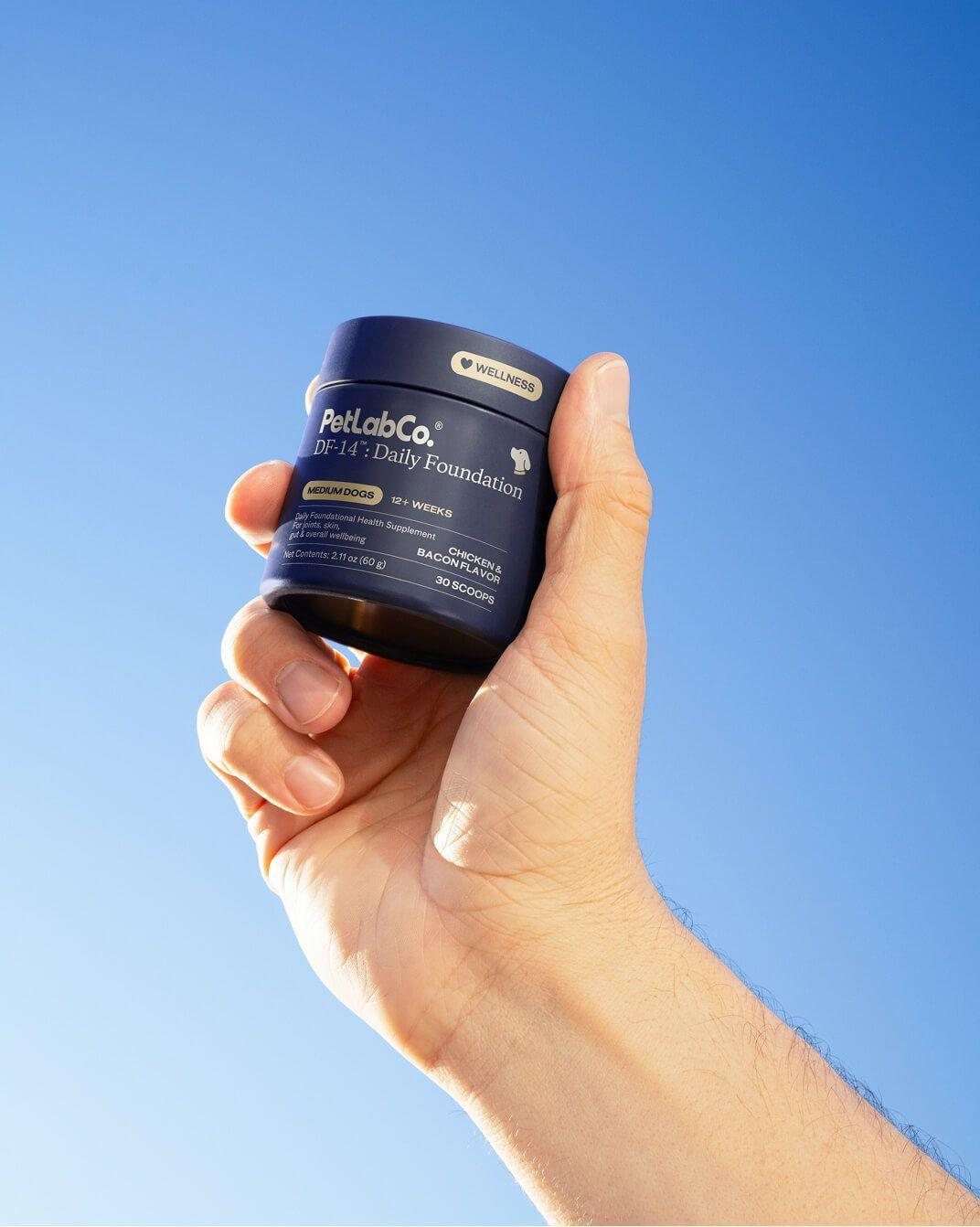 A hand holds a DF-14 Daily Foundation tub against a blue sky.